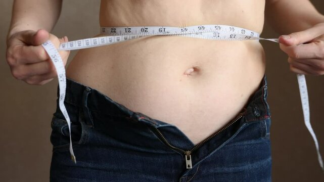 Fat woman in unbuttoned jeans measures her waist with tape measure and then desperately squeezes her belly fat with tape measure, close-up. Lifestyle, overweight concept, weight loss, self-loathing.