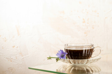 Glass cup of chicory tea on saucer with lilac flower on glass table near textured wall. Copy space.