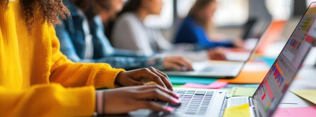 Close-up of young people working together on a laptop computer with colorful papers and pens at an office desk.