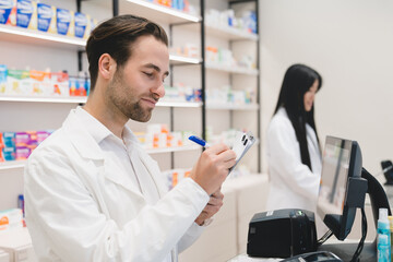 Two young pharmacists druggists working together at cash point desk in drugstore pharmacy using computer and writing on clipboard