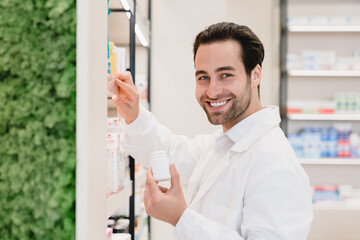 Male young caucasian handsome pharmacist druggist in white medical coat holding remedy pills while looking for another medicines on shelves of pharmacy drugstore looking at camera