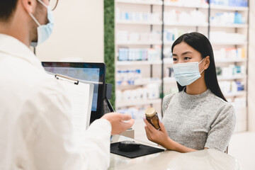 Male pharmacist druggist in protective face mask against coronavirus advising female customer client buyer remedy, medicines, pills, antibiotics against Covid19 in pharmacy