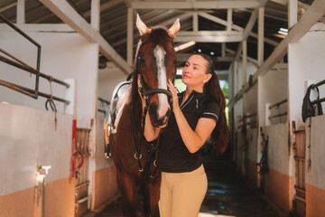 Young woman rider in equestrian uniform standing in stable next to her horse and petting it....
