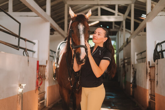 Young woman rider in equestrian uniform standing in stable next to her horse and petting it. Horseback riding training. Love and affection for domestic brown horse. Authentic village barn. - Powered by Adobe