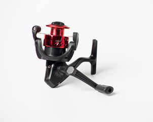 A red and black fishing reel sits on a white background. The reel is small and has a black handle
