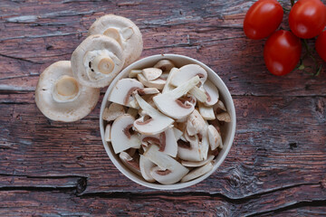 A top view of a bowl filled with freshly sliced mushrooms, placed on a rustic wooden table. Whole mushrooms and ripe tomatoes are arranged nearby, showcasing fresh ingredients for meal preparation..