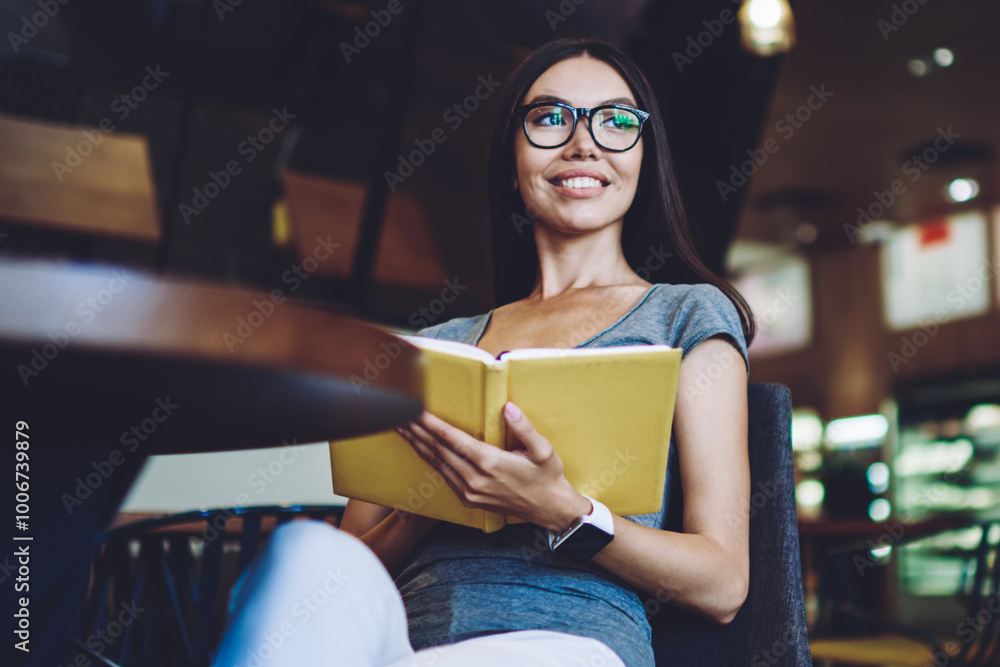 Poster cheerful hipster girl with notepad in hands smiling and looking aside while spending time for litera