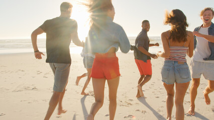 Rear View Of Casually Dressed Friends Having Fun Running Along Summer Beach Together With Lens Flare
