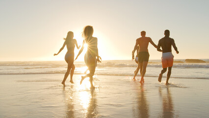 Fototapeta premium Group Of Friends In Swimwear Run Through Waves At Sunset On Summer Beach Vacation With Lens Flare