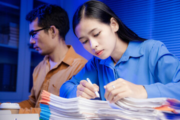 Two Asian women work late into the night. They are exhausted and stressed at their desks. They are...