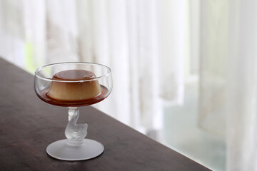 Caramel flan or crème caramel, elegantly served in a glass dessert cup. The cup has a delicate, artistic stem that resembles a swan. The flan sits in a pool of caramel sauce,with smooth glossy texture