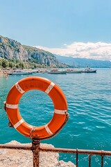 An orange lifebuoy with white stripes is mounted on a weathered metal pole on a stone pier overlooking a scenic bay. 