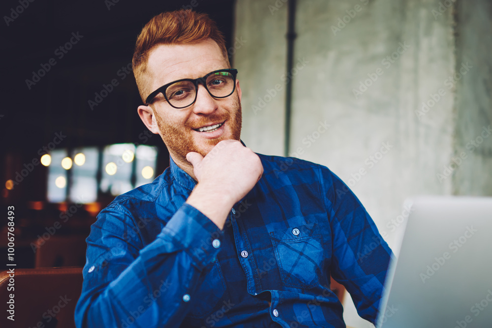 Poster portrait of cheerful male freelancer in stylish eyeglasses looking at camera while sitting with lapt
