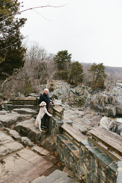 Older man hikes in mountain and rocks with tree and his pet dog