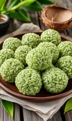 Obraz premium Green sticky rice balls on a brown plate with a coconut half shell, green leaves, and a wooden table background.