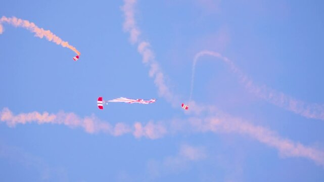 British Red Devils parachutists descend with precision, leaving vibrant red and white smoke trails in the sky during a military display. Captured mid-air against a clear blue backdrop.