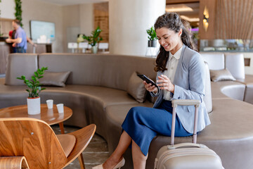 A smiling businesswoman sits in a modern hotel lobby, checking her phone while holding onto her...