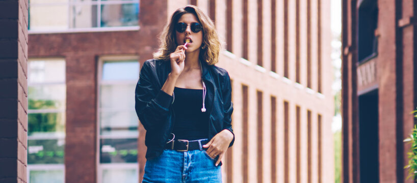Full length portrait of confident female traveller posing with chupa chups on publicity area, attractive caucasian hipster girl in sunglasses looking at camera during vacations in other city