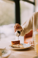 A joyful moment at a cozy cafe as a woman pours syrup over a slice of cheesecake, capturing the essence of indulgence and relaxation