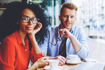 Portrait of positive multicultural male and female friends smiling at camera while spending leisure time togetherness for download movie on laptop computer with good internet connection in cafeteria