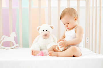 a little baby girl with a bottle or a drinking cup in her hands in a crib at home in a children's room in a white bodysuit smiling or laughing, cute funny baby, lifestyle