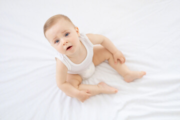 happy little baby girl or boy in a white bodice on the bed in the bedroom looking up, baby with blue eyes