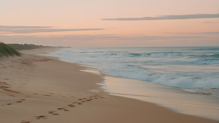 Obraz premium Footprints in the sand along a serene beach with gentle waves rolling in at sunrise, casting soft pastel hues across the sky and ocean