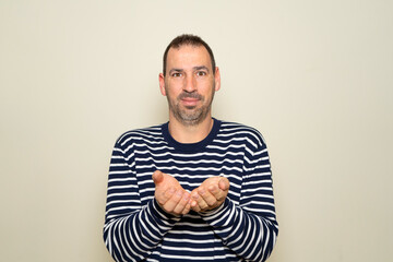 Fototapeta premium Bearded hispanic man in his 40s wearing a striped sweater offering something imaginary on the palms of his hands, exuding kindness and hospitality. Isolated over beige background.