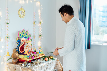 Indian man dressed in traditional wear engages in worship at home altar, adorned with decors and offerings for god Ganesh during festive celebration. Indian culture, hindu ritual and customs.