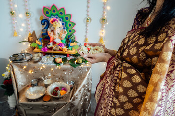 Indian woman dressed in traditional wear engages in worship at home altar, adorned with decors and...