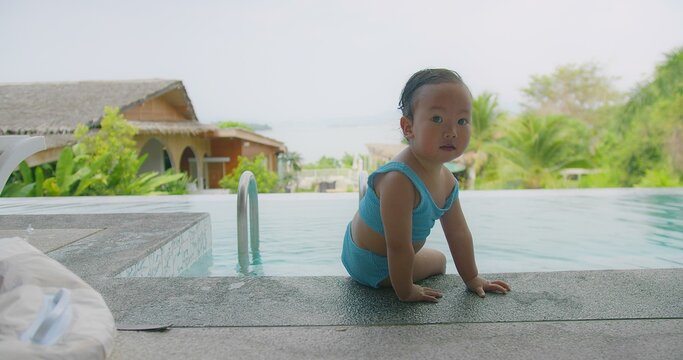 Cute little child girl in a blue swimsuit sitting at poolside looking smiling at camera surrounded by tropical greenery and resort buildings, enjoying a sunny day.