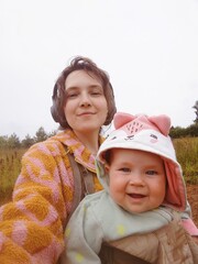 A young mother and daughter. Parent hiking with her child in baby carrier. Taking care of 9 month old baby. Active parenting close to nature. Woman listening to music in headphones.