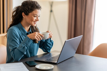 A woman in a blue shirt sits at a desk with a laptop, eyes closed, smiling, and enjoying a cup of...