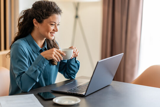 A woman in a blue shirt sits at a desk with a laptop, eyes closed, smiling, and enjoying a cup of coffee. She looks relaxed, taking a break during work in a calm setting.