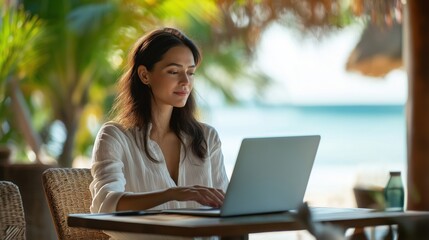 A young woman sitting at a table, working on her laptop by the beach with palm trees in the background.