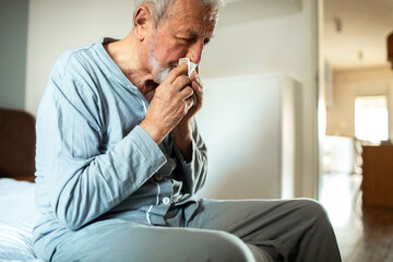 Senior man in pajamas sneezing into tissue while sitting on bed