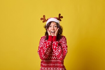 Excited young woman in red holiday sweater with snowflake and reindeer pattern, wearing reindeer antler headband, hands raised to face in surprise. Isolated on yellow background.