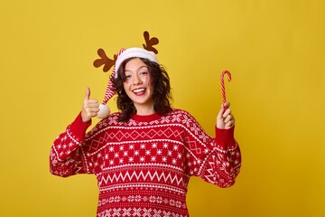 Expressive curly-haired female model in red knitted sweater with white snowflakes pattern and reindeer antlers headband gesturing thumbs up and holding candy cane on yellow background