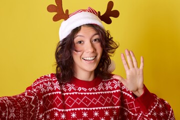 Young woman with brown hair and warm smile wearing a red Santa hat, reindeer antlers, and cozy...