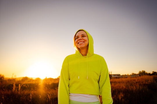 A joyful woman in a bright hoodie enjoys a sunset in a golden field while smiling and embracing the moment