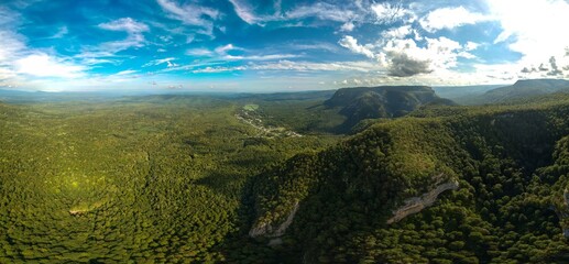 aerial panorama of mountains covered with green forest, small village Guamka (Caucasus, Russia) in the distance, mountain slope with grottoes on a sunny summer day
