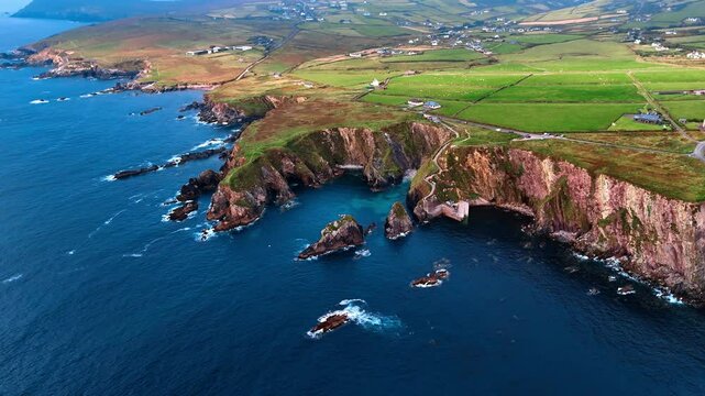 Deep blue waters of the Atlantic Ocean splash by the stunning rocks at the shore. Beautiful scenery of the coastline of Ireland from drone. Multiple cottages are in the valley among the mountains.