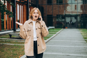 Fototapeta premium Angry woman on cell phone walking outside building. Frustrated woman talking on phone. Summer time. Negative people emotion
