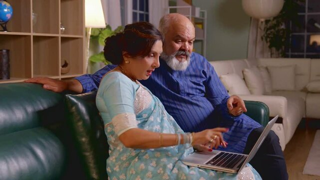 Indian Asian old couple sitting on a sofa at home, working together on a laptop, sharing ideas and enjoying their retired life