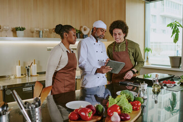 Professional chef holding touchpad with new menu and discussing it with his assistants while they standing in center of kitchen