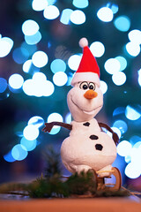 Toy cute snowman on the sleigh against the background of blurred lights garlands.