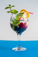 cocktail raspberry blueberry lemon and mint in a glass with a straw on a blue background.