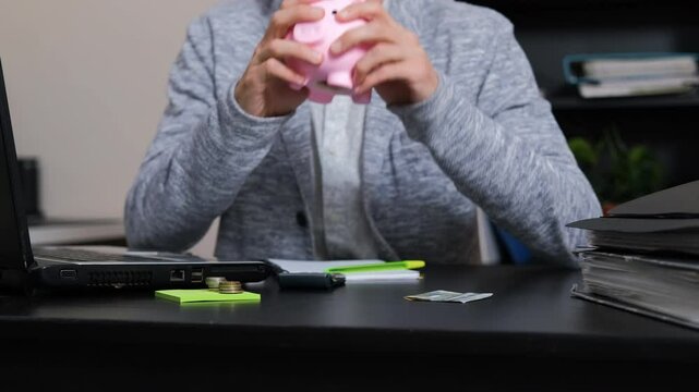 Man sits at his office desk, eagerly emptying a pink piggy bank in the office setting. Coins scattering across the surface, slow motion. Savings, budgeting and financial planning concept