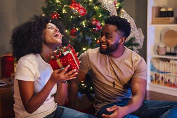 African american woman joyfully receives gift from her partner by Christmas tree