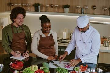 High angle shot of chef in white uniform cutting red onion while his biracial interns writing down his actions